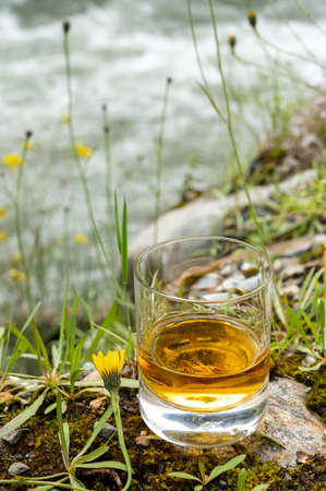 Glass of strong scotch single malt whiskey with fast flowing mountain river and wild flowers on...