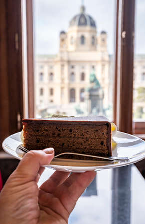 Hand with piece of famous Sachertorte chocolate cake with apricot jam of Austrian origin and window view on old Viennaの写真素材
