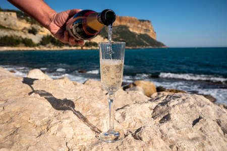 Pouring of French champagne sparkling wine in glass and view on beach, blue sea water and mountains...