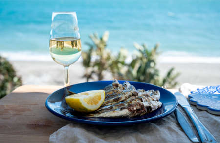 Sardines espeto prepared on skewers and open flame on fireplace with olive trees wood, served outdoor with lemon and glass of fino sherry wine and view on blue sea, Malaga, Andalusia, summer vacation in Spainの写真素材