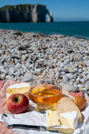 Lunch on pebbles beach of Etretat, french cheese camembert, fresh baked baguette and apple cider drink with chalk cliffs and Atlantic ocean on background, Normandy, Franceの写真素材