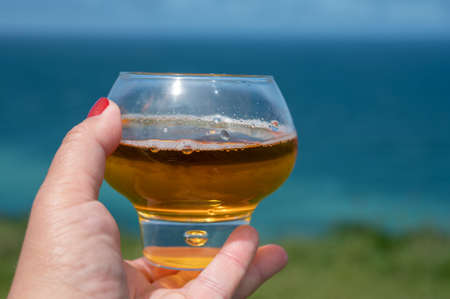 Woman's hand with glass of apple cider drink and view on on green grass fields on chalk cliffs of...