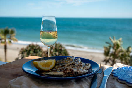 Sardines espeto prepared on skewers and open flame on fireplace with olive trees wood, served outdoor with lemon and glass of fino sherry wine and view on blue sea, Malaga, Andalusia, summer vacation in Spainの写真素材