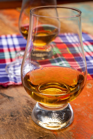 Tulip-shaped tasting glass with dram of Scotch single malt or blended whiskey on wooden table with Scottish tartanの写真素材
