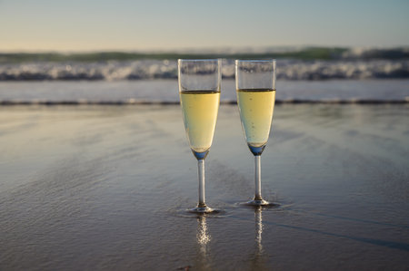 Glasses of cava or champagne sparkling wine on white sandy ocean beach with water waves on sunset in sunlights with shadowsの写真素材