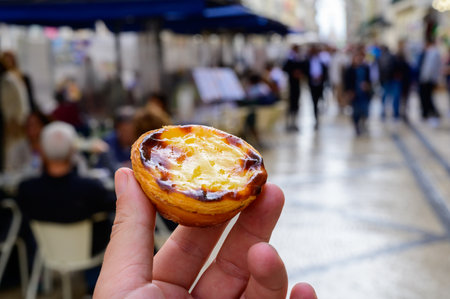 Hand with Portugal's traditional sweet dessert Pastel de nata egg custard tart pastry and view on old street in Lisbon, Portugalの写真素材