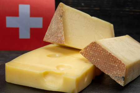 Assortment of Swiss cheeses Emmental or Emmentaler medium-hard cheese with round holes, Gruyere, appenzeller used for traditional cheese fondue and gratin and flag of Switzerland on dark backgroundの写真素材
