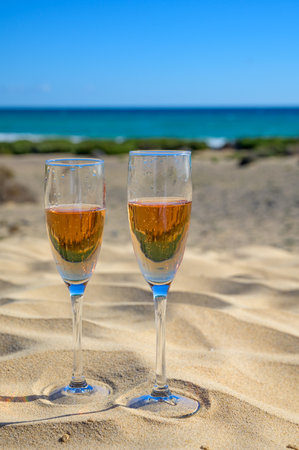 Two glasses of rose champagne or cava sparkling wine served on the white sandy tropical beach with dunes and blue ocean, romantic vacation, winter sun on Fuerteventura, Canary, Spainの写真素材
