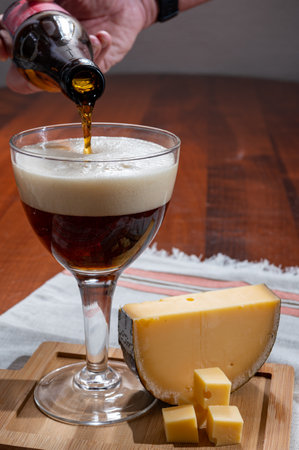 Pouring of Belgian abbey brown beer in glass and wooden board with old belgian cheese made by monks from cow milk, tastes of Belgiumの写真素材
