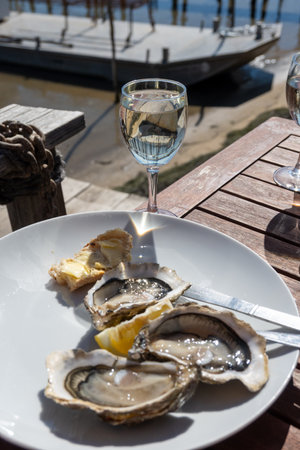 Eating of fresh live oysters with glass of white wine at farm cafe in oyster-farming village, with view on boats and water of Arcachon bay, Cap Ferret peninsula, Bordeaux, France in sunny dayの写真素材