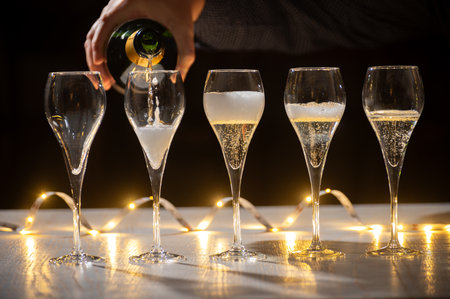 New year party, pouring of brut champagne bubbles cava or prosecco wine in tulip glasses with garland lights on dark backgroundの写真素材