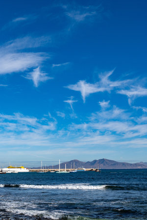 Walking on sea promenade in Corralejo along black rocks, blue water, view on volcanic Lanzarote island, Fuerteventura, Canary islands, Spainの写真素材