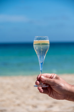 Hand with glass of cava or champagne sparkling wine on vacation, Dunes Corralejo Grand sandy beach, Fuerteventura, Canary islands, blue oceanの写真素材