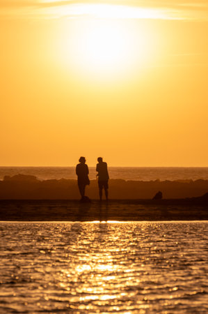 Man and woman staying and looking at sunset on ocean beach, orange sky, silhouettes of people on vacation, Canary islandsの写真素材