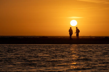 Man and woman staying and looking at sunset on ocean beach, orange sky, silhouettes of people on vacation, Canary islandsの写真素材