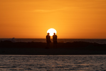 Man and woman staying and looking at sunset on ocean beach, orange sky, silhouettes of people on vacation, Canary islandsの写真素材