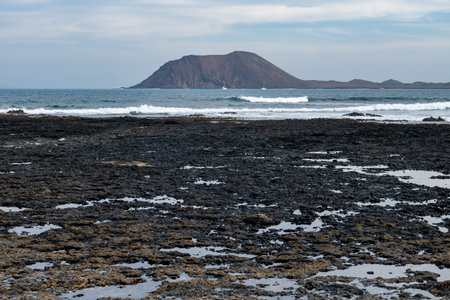 Walking on sea promenade in Corralejo along black rocks, blue water, view on volcanic Lobos island, Fuerteventura, Canary islands, Spainの写真素材