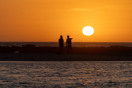 Man and woman staying and looking at sunset on ocean beach, orange sky, silhouettes of people on vacation, Canary islandsの写真素材