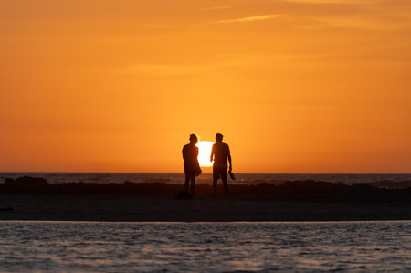 Man and woman staying and looking at sunset on ocean beach, orange sky, silhouettes of people on vacation, Canary islandsの写真素材