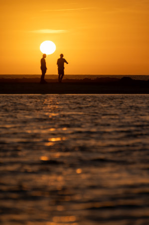 Man and woman staying and looking at sunset on ocean beach, orange sky, silhouettes of people on vacation, Canary islandsの写真素材