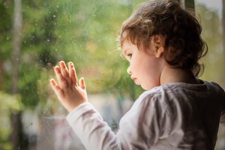 Photo of a little curly girl on the window sill, it is raining outside the window.の写真素材