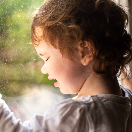 Photo of a little curly girl on the window sill, it is raining outside the window.の写真素材