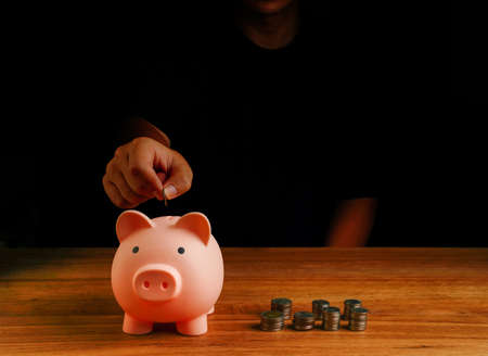 Hand put coin into piggy bank, saving cash coin concept investment. Management growth funding budget for wealth. Wooden background with saving word and coins on it.の写真素材