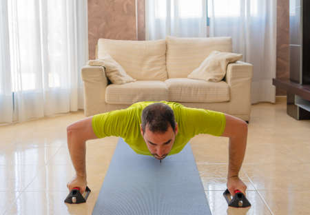 Bearded man in low shape exercising with black and green sportswear in his living room in front of...