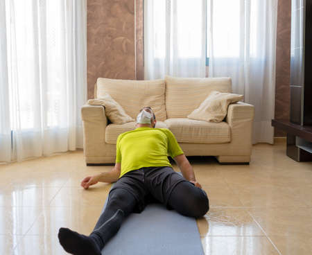 Bearded man in low shape exercising with black and green sportswear and a mask to prevent...