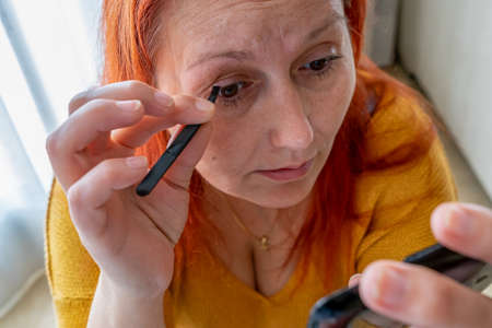 Beautiful redhead woman in a yellow sweater plucking her eyebrows on living room at homeの写真素材