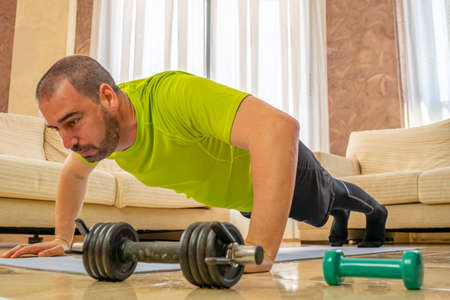 Bearded man in low shape exercising with black and green sportswear in his living room in front of...