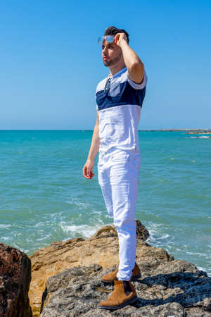 Young man in white pants and modern styling posing on the beach of the Mediterranean Sea in...