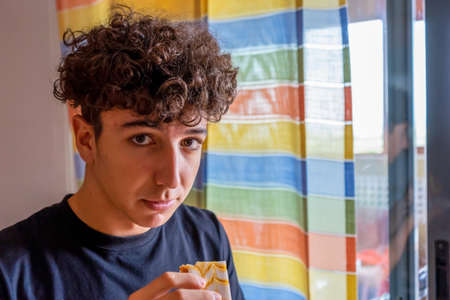 Attractive young man with curly hair eating a sweet in the kitchen of his home. Food conceptの写真素材