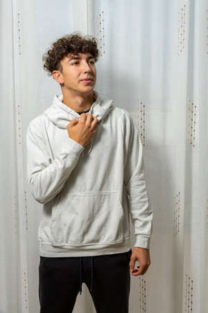 Attractive young man with curly hair wearing white sweatshirt posing on white curtains background....