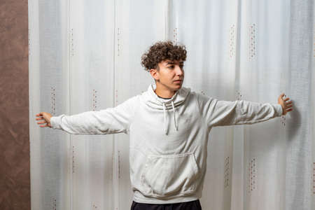 Attractive young man with curly hair wearing white sweatshirt posing on white curtains background. Fashion conceptの写真素材