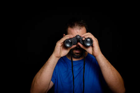 Bearded man dressed in blue t-shirt with binoculars posing against black background. Fashion concept