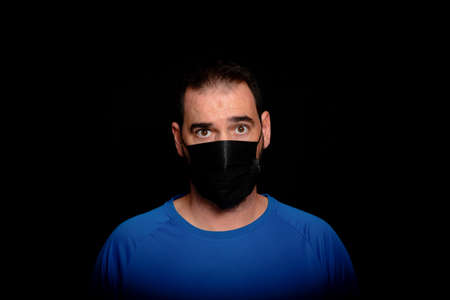 Bearded man dressed in blue t-shirt and with a black mask to protect himself from coronavirus posing against black background. Health conceptの写真素材