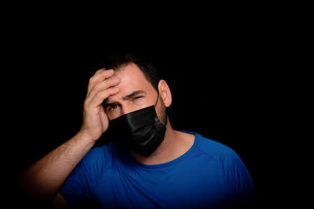 Bearded man dressed in blue t-shirt and with a black mask to protect himself from coronavirus posing against black background. Health conceptの写真素材