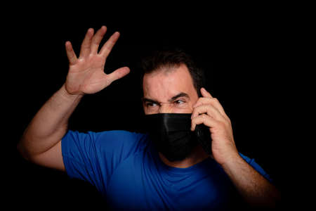 Bearded man dressed in blue t-shirt and with a black mask to protect himself from coronavirus posing against black background. Health conceptの写真素材