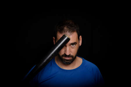 Bearded man dressed in blue t-shirt in defiant pose posing with a baseball bat on  blackの写真素材