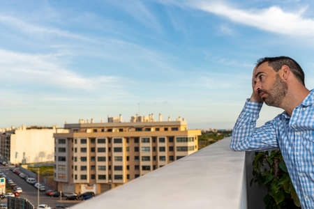 Young attractive, sad and desperate hispanic man suffering from depression who looks thoughtful and frustrated on the terrace of his home looking depressed to the street. Depression conceptの写真素材