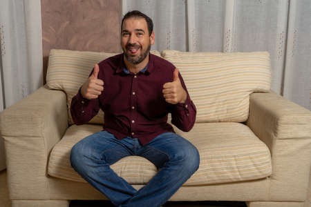 Bearded man with dental braces sitting in armchair with thumbs up sticking out his tongue. Happiness conceptの写真素材