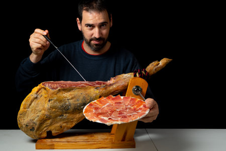 Bearded man with a ham knife ready to cut a piece of typical Spanish ham isolated on black studio background. Super foods. Delicious appetizer. Feeding conceptの写真素材