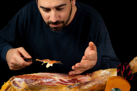 Bearded man with a ham knife ready to cut a piece of typical Spanish ham isolated on black studio background. Super foods. Delicious appetizer. Feeding conceptの写真素材