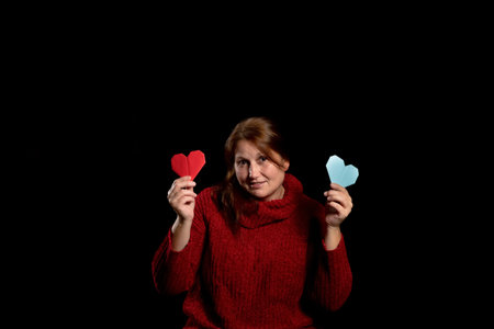 Pretty woman in a red woolen sweater with a red heart and blue one made of paper posing on black studio background. Copy space. Valentine's Day. Love conceptの写真素材