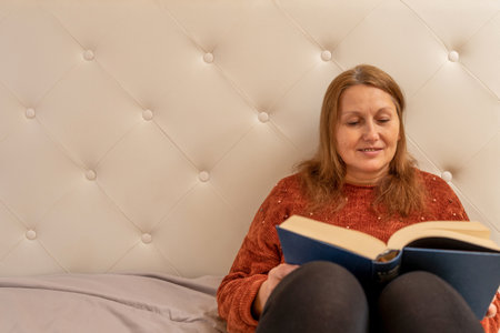 Pretty adult woman reading a book sitting on the bed in her room. Entertainment concept. Selective focusの写真素材