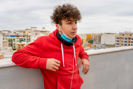 Young attractive man with curly hair dressed in a red sweatshirt using mobile on the terrace of the...