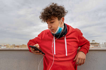 Young attractive man with curly hair dressed in a red sweatshirt using mobile on the terrace of the...