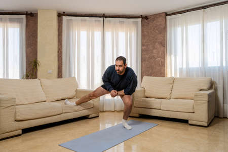 Fit bearded man in black sportswear doing stretching exercise in living room at home. Training...