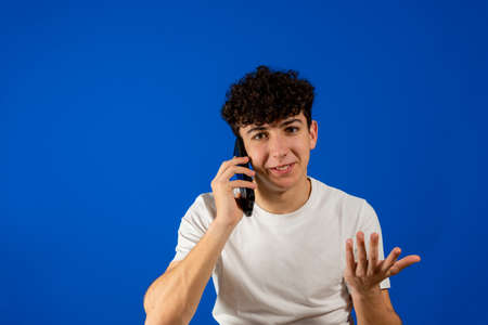 Handsome young man with curly hair talking on the smartphone isolated on blue studio background. Technology conceptの写真素材